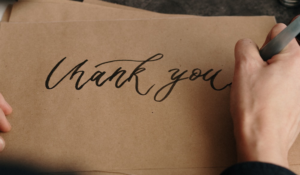 A hand writing "thank you" on a pica of brown paper.
